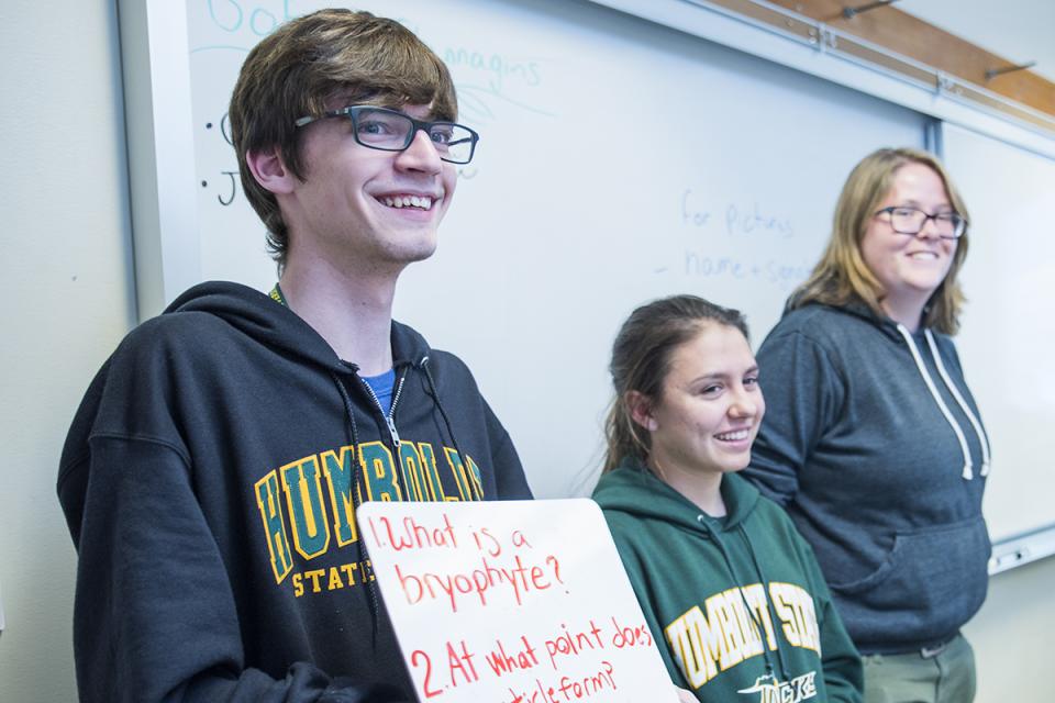 Three students standing in front of a class presenting a sign