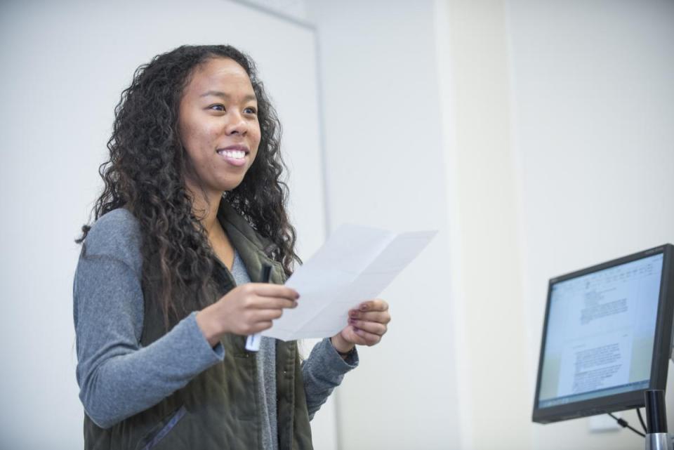 A woman standing in front of a class smiling and holding a piece of paper